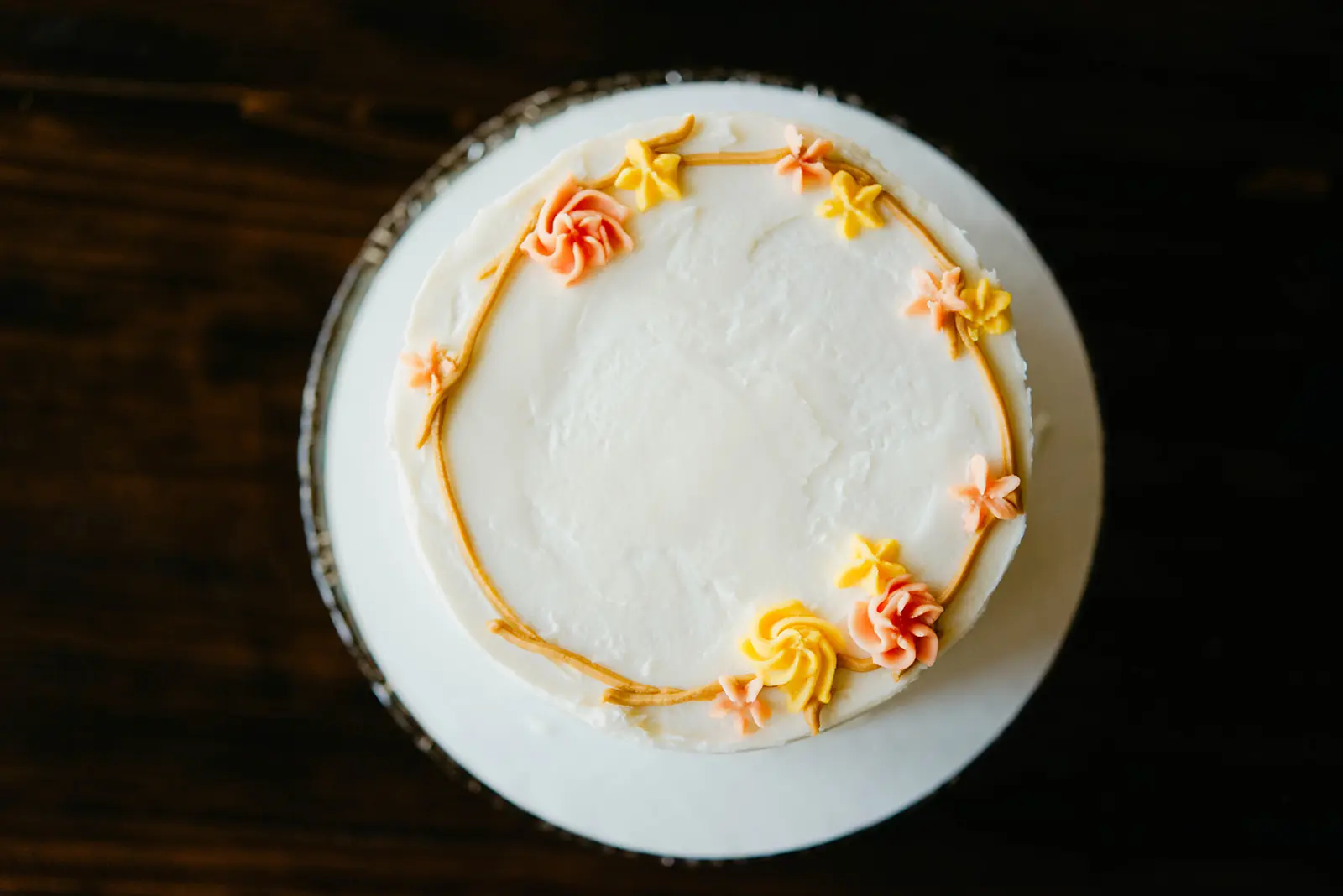 Overhead view of a white cake with piped peach flowers on a pedestal against dark wood
