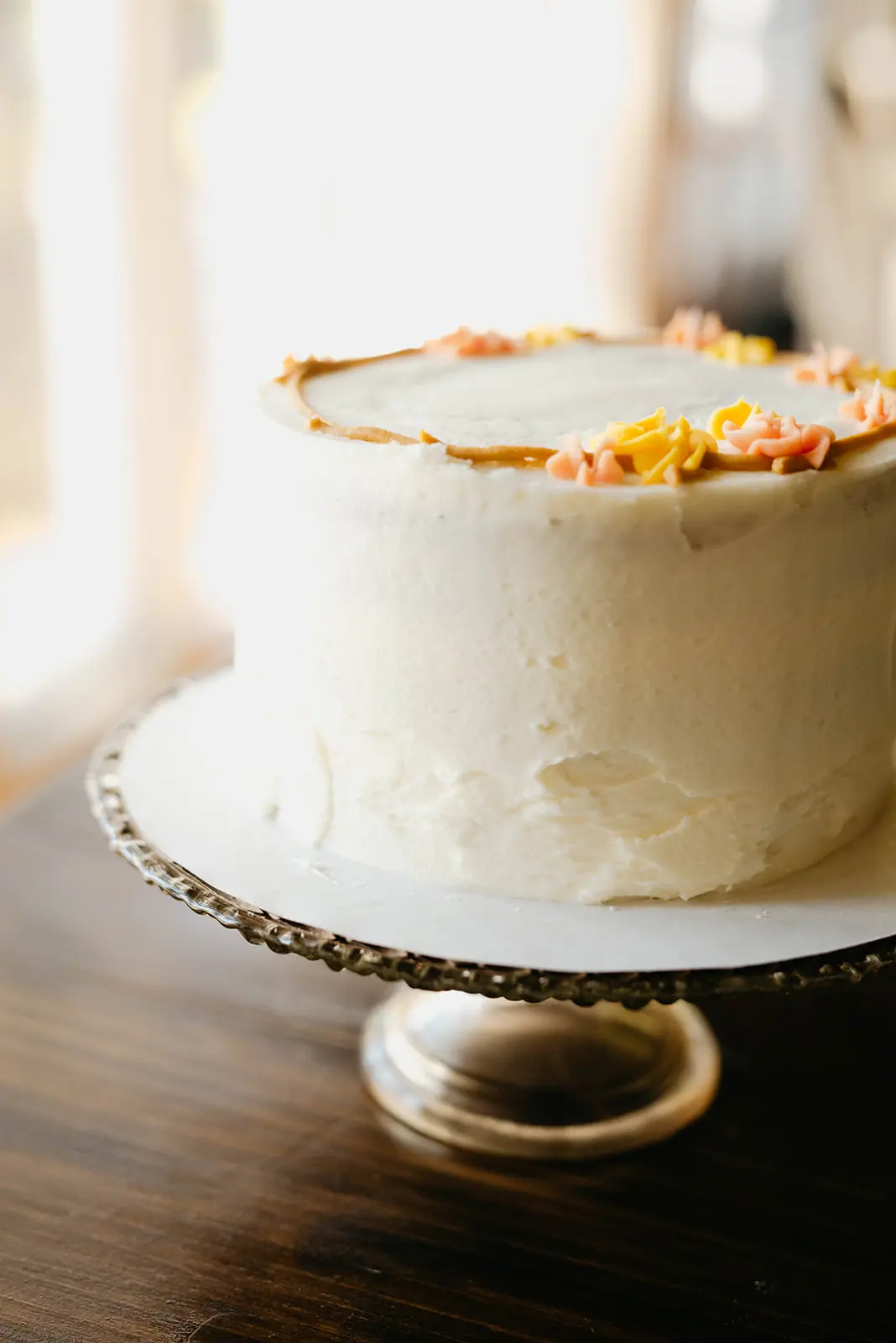 A white buttercream cake with piped flowers on a glass pedestal in bright window light