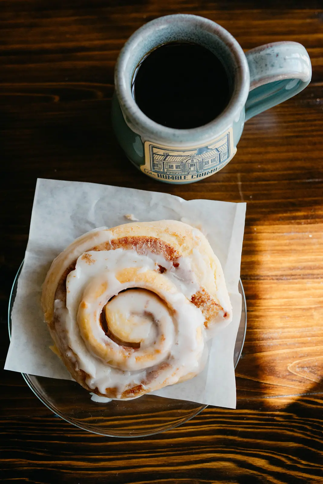 A glazed Humble Crumb cinnamon roll beside coffee in a branded bakery mug