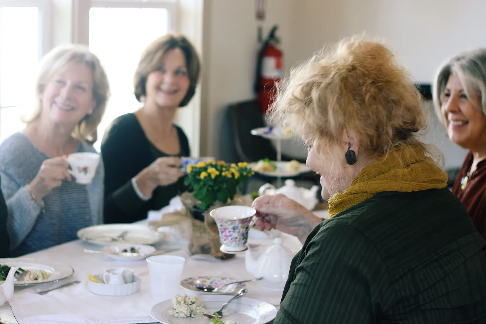 Four guests drinking tea at a white-linen table with floral cups and a teapot