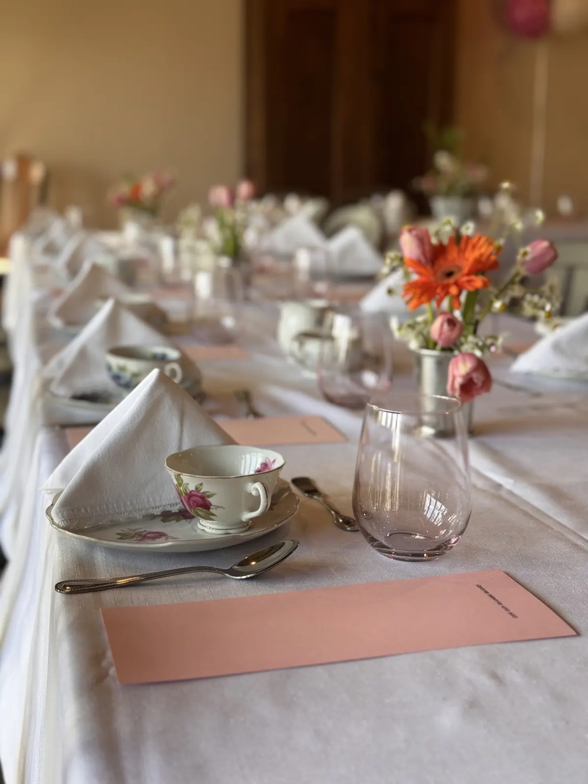 A long tea table with rose-patterned cups, folded napkins, and orange and pink flowers